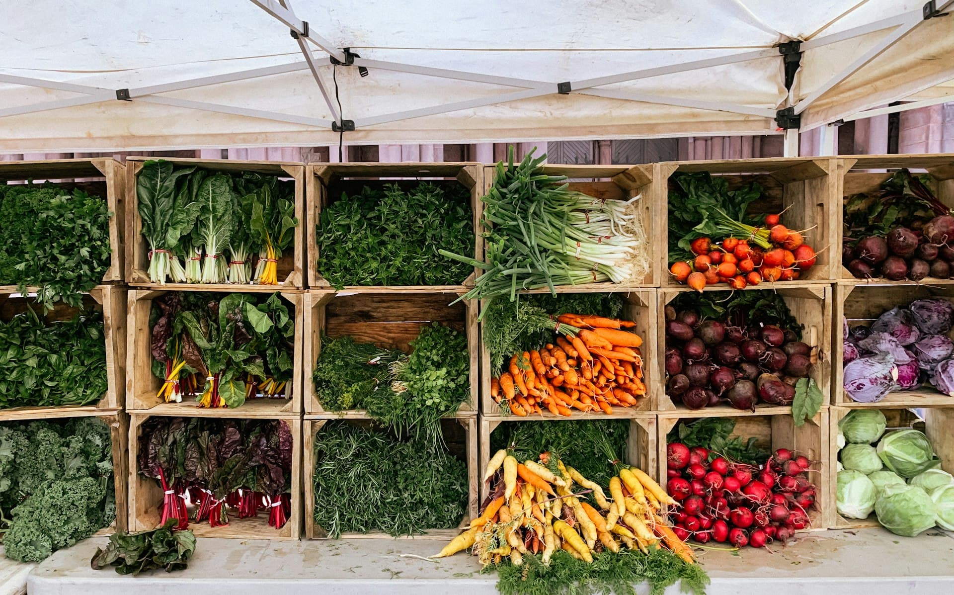 Colorful fresh vegetables displayed in wooden crates at a farmers market stand, including green leafy vegetables, orange carrots, red tomatoes, and purple beets under a canvas canopy.