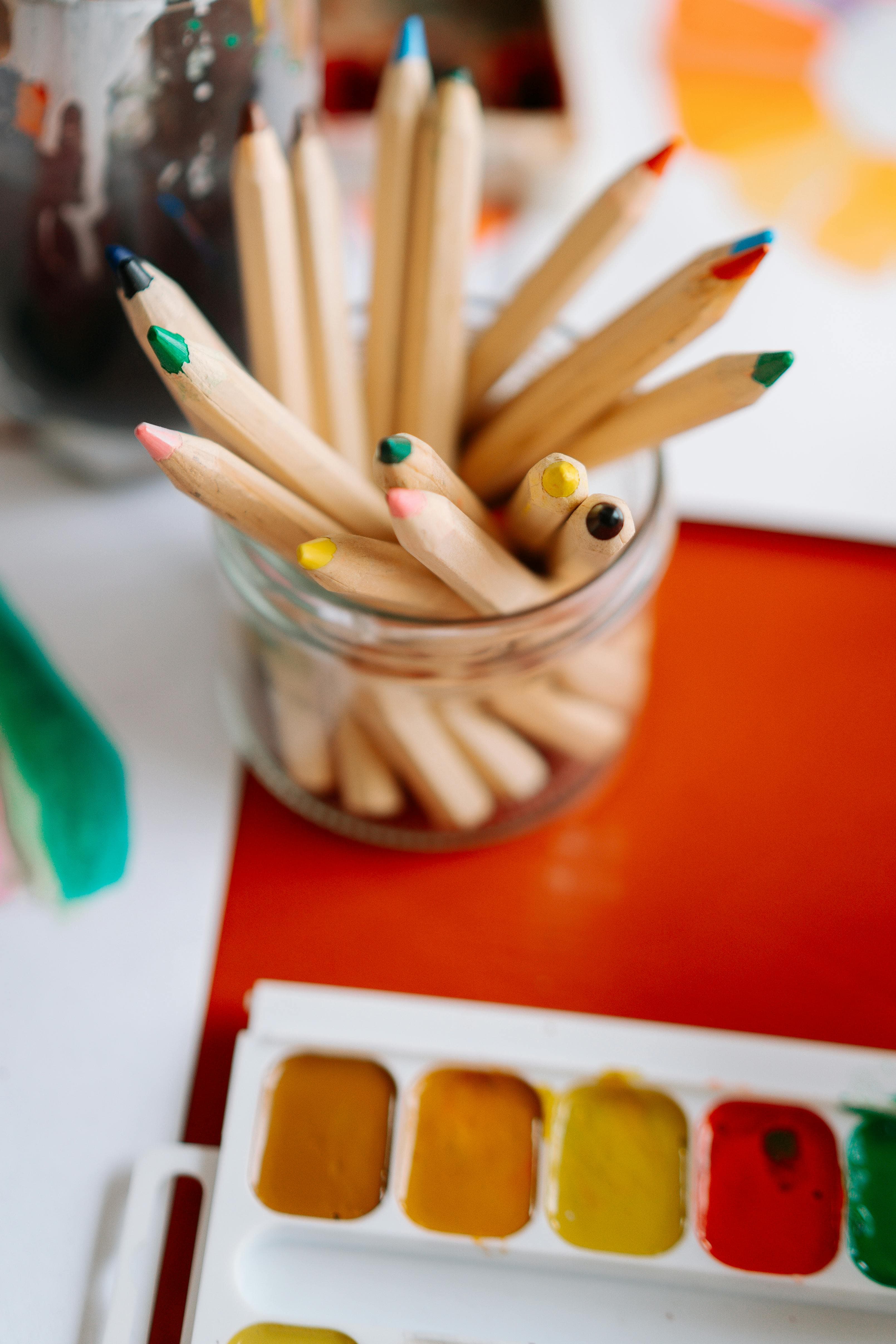 Jar of thick colored pencils and markers beside a watercolor palette
