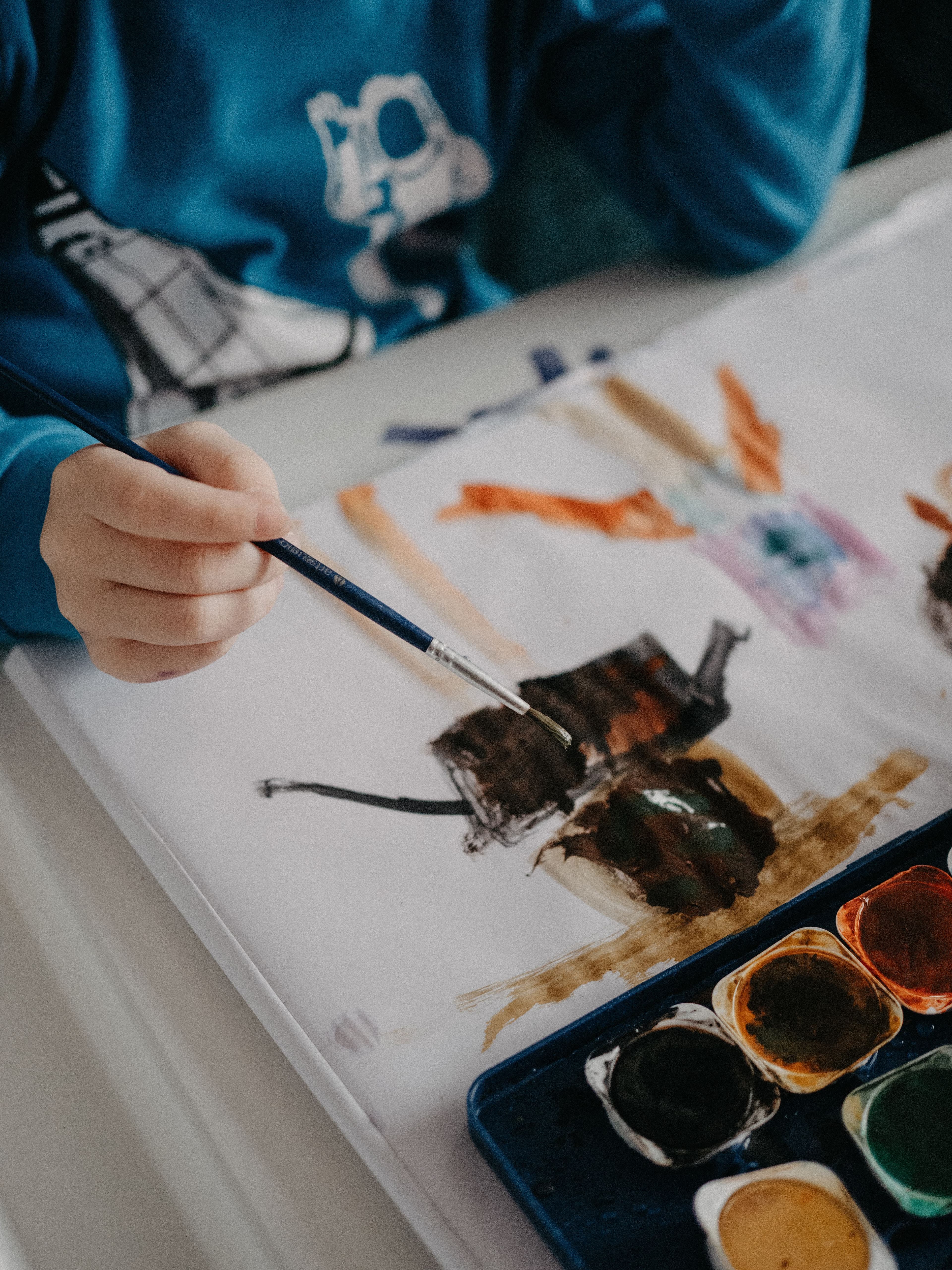 Child painting with watercolors on open paper beside a palette