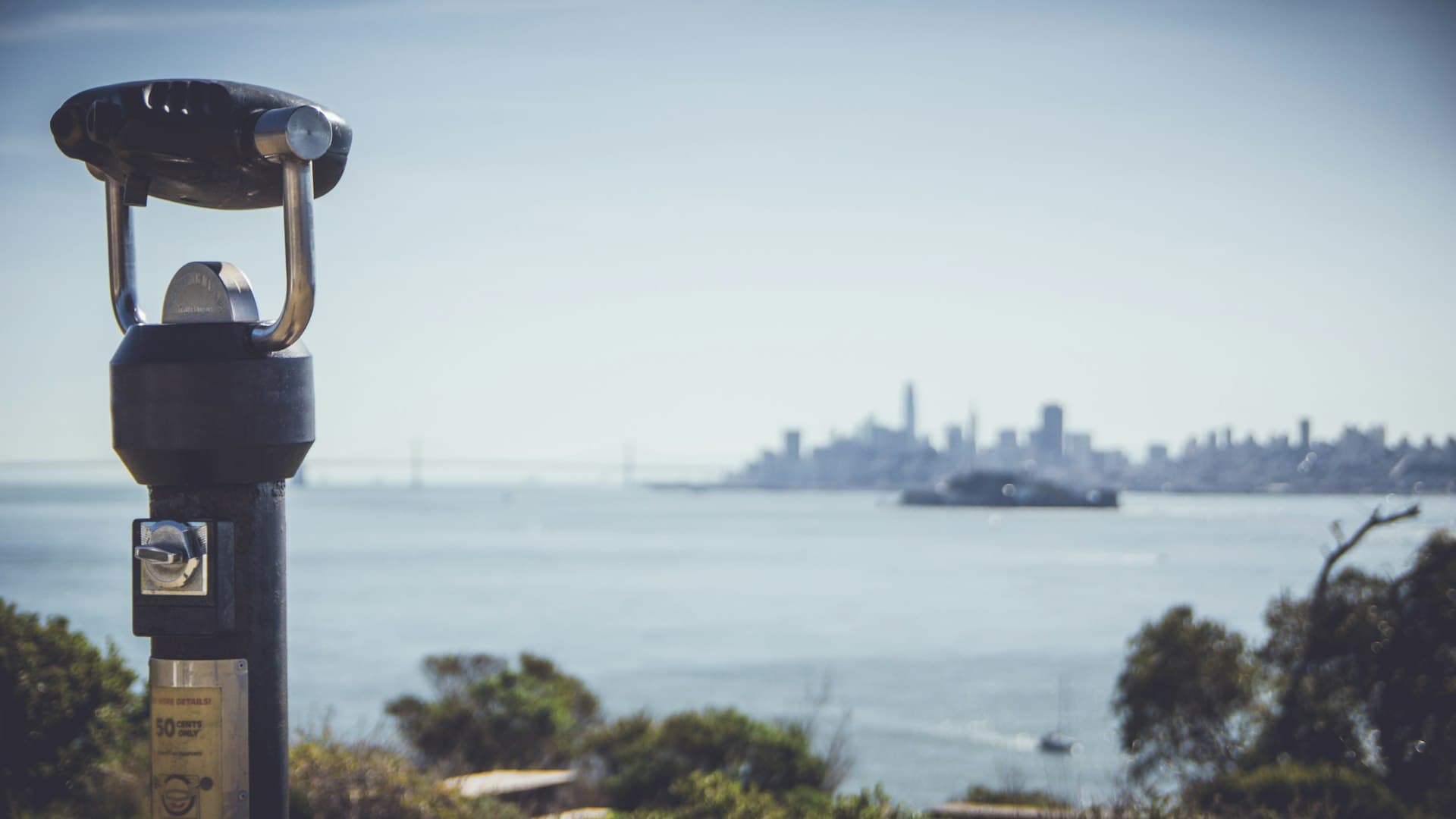A public telescope on a waterfront overlook pointing toward a distant city skyline across calm water on a clear day.