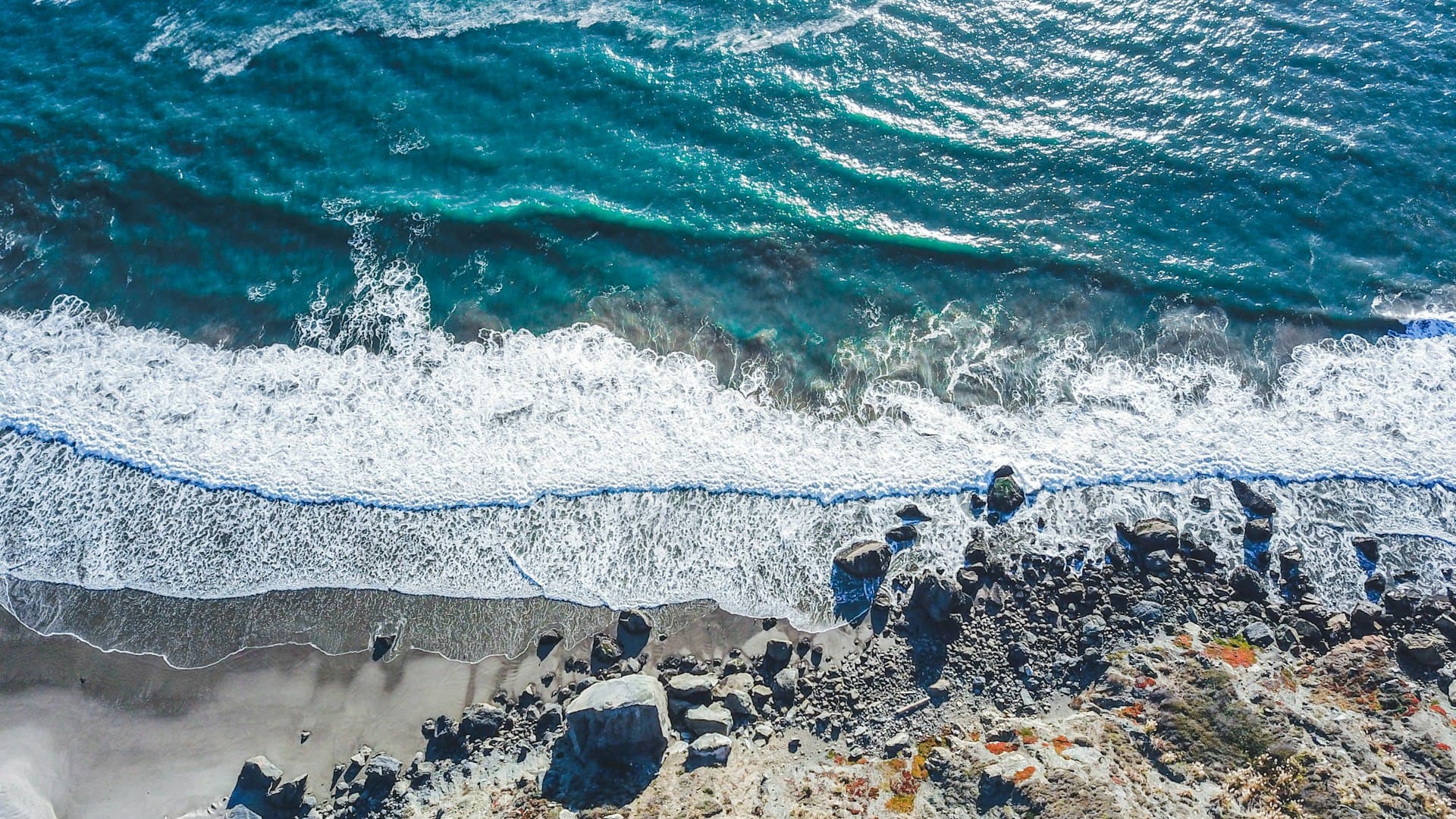 Aerial view of waves crashing on a rocky beach with turquoise ocean water and dark boulders