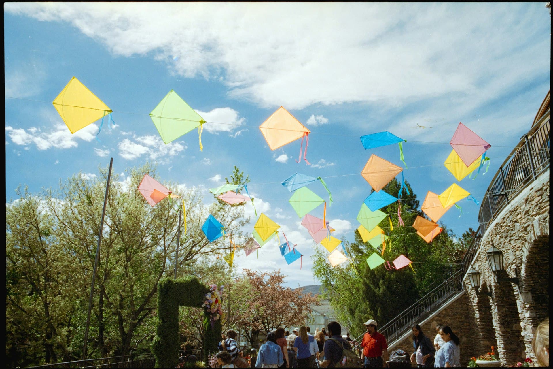 Children and families flying colorful diamond kites in a sunny outdoor plaza with historic stone buildings and green trees in the background.