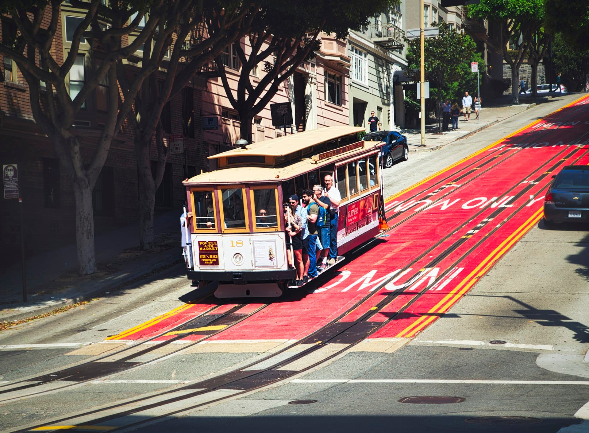 A multi-generational family rides a cable car down a steep San Francisco street with historic buildings and trees in the background on a sunny day.
