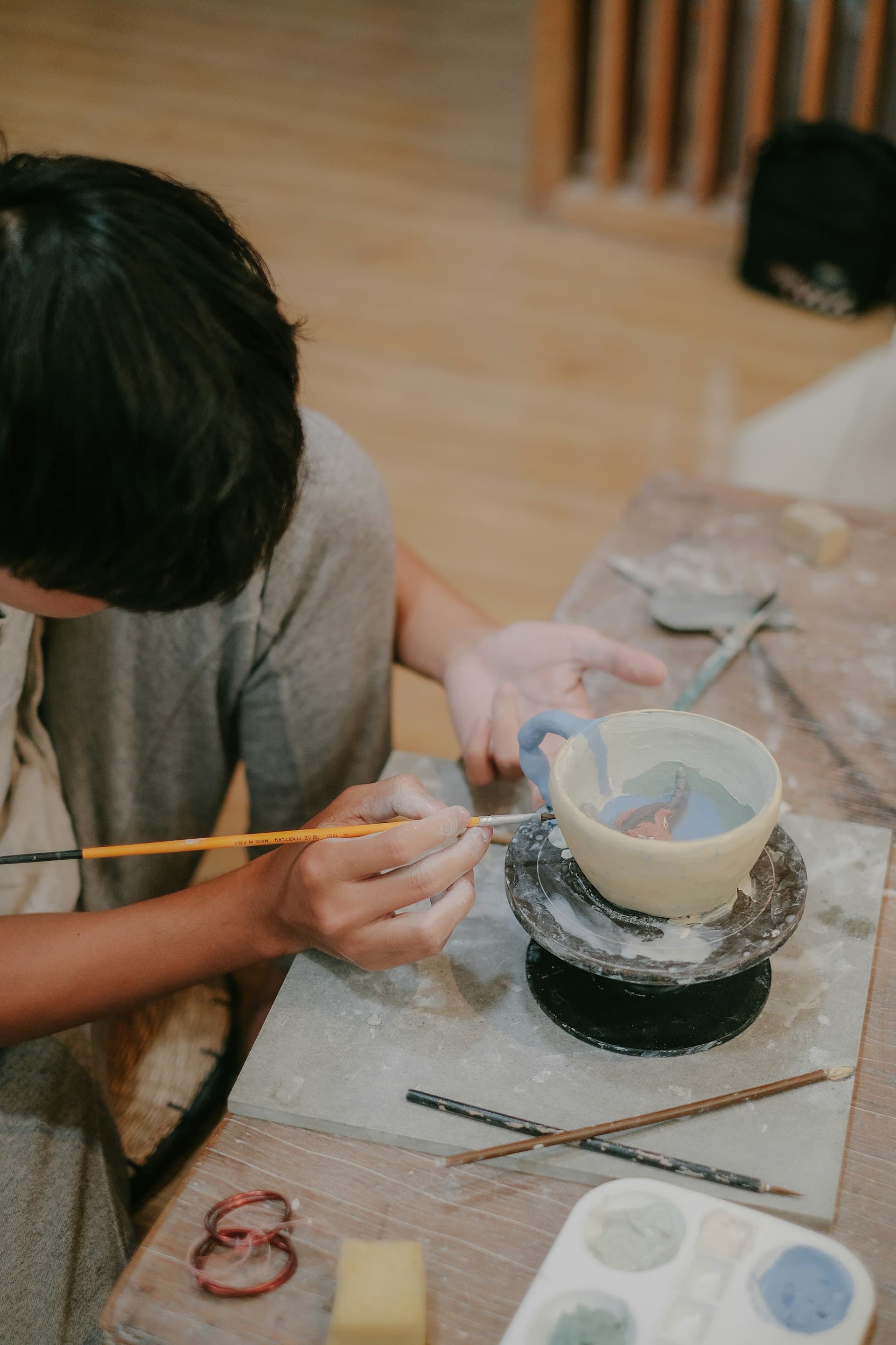 A school-age child painting a ceramic bowl with a brush while seated at a wooden craft table with art supplies.