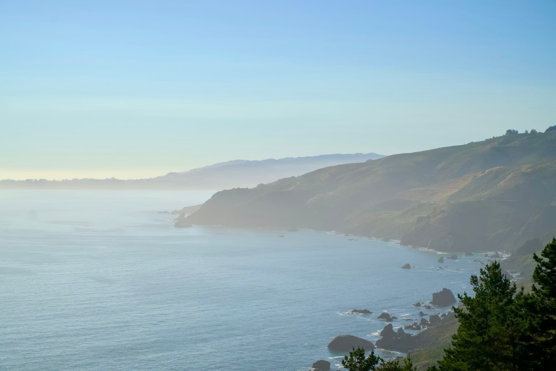 Panoramic view of misty coastal cliffs overlooking a calm ocean at golden hour, with layered mountains and tree-covered headlands creating a serene natural landscape.