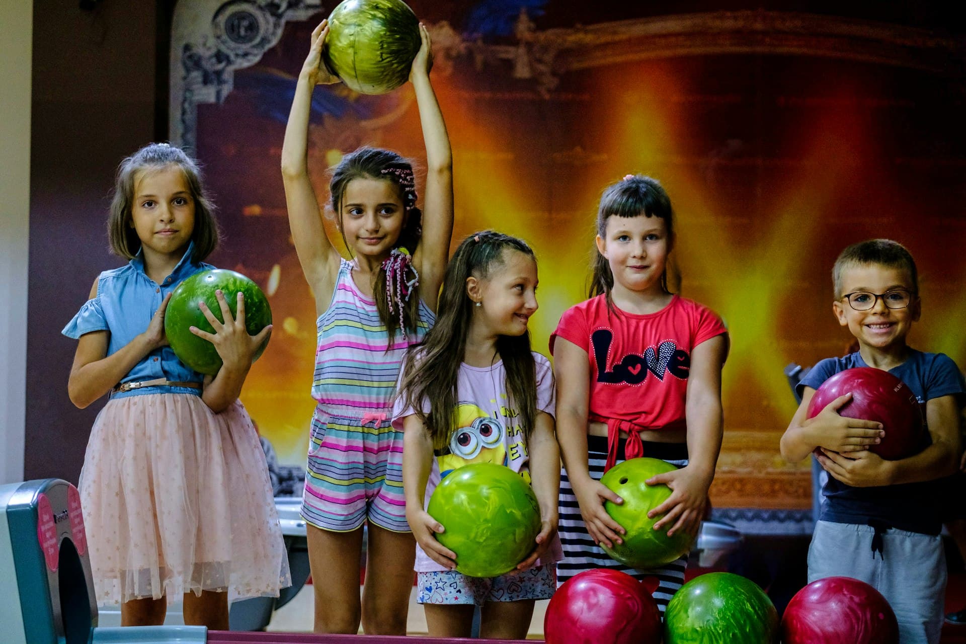 Five smiling children holding colorful bowling balls at a bowling alley with warm lighting in the background.