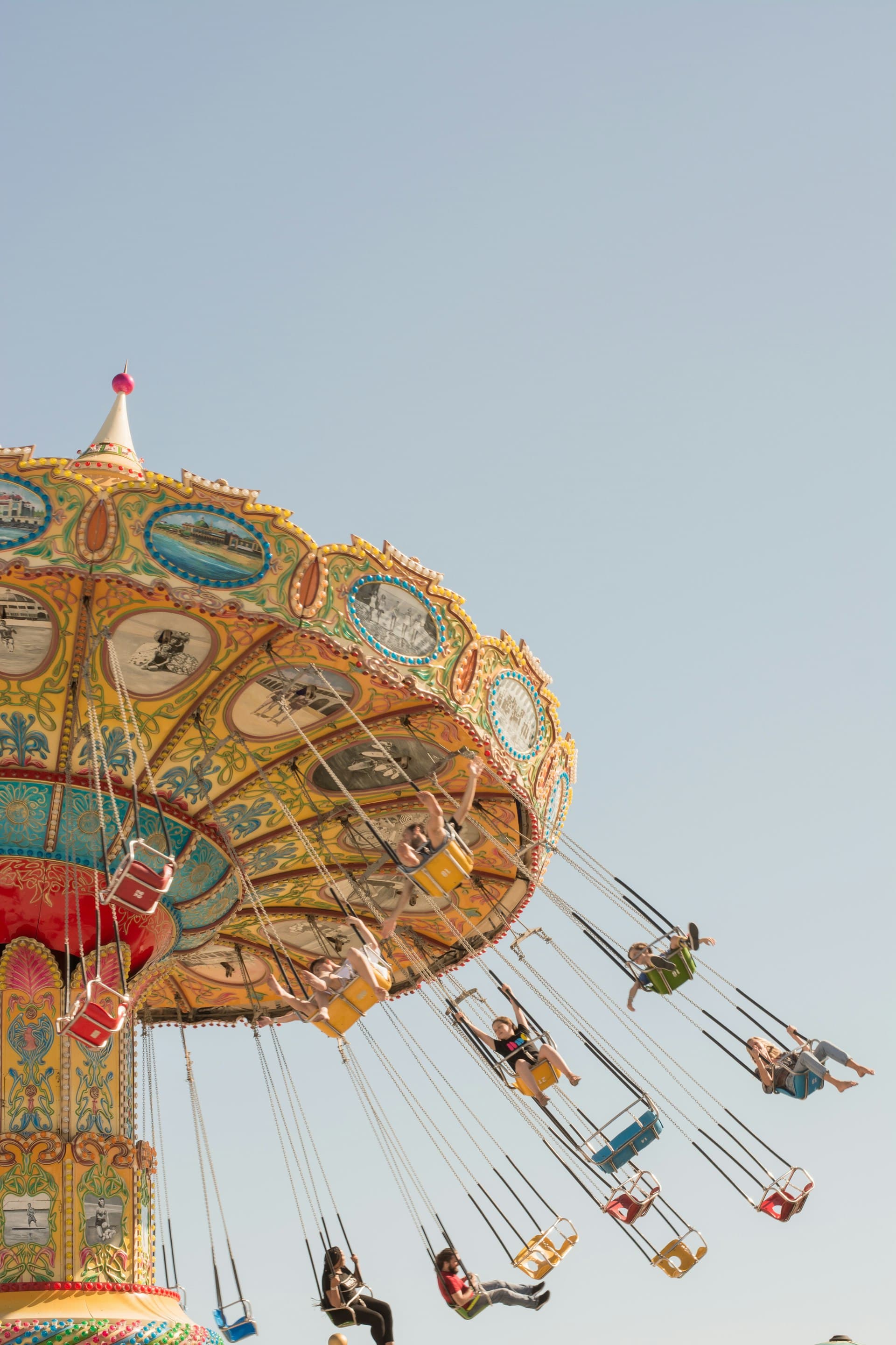 Families riding a colorful ornate carousel swing ride against a blue sky at an amusement park