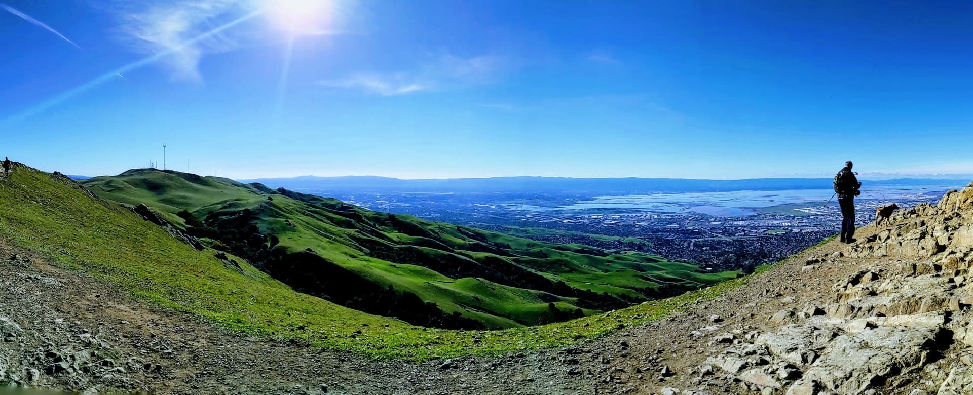 Solitary hiker in backpack standing on a rocky ridge overlooking a vast panorama of green rolling hills and distant valley under bright sunny sky.