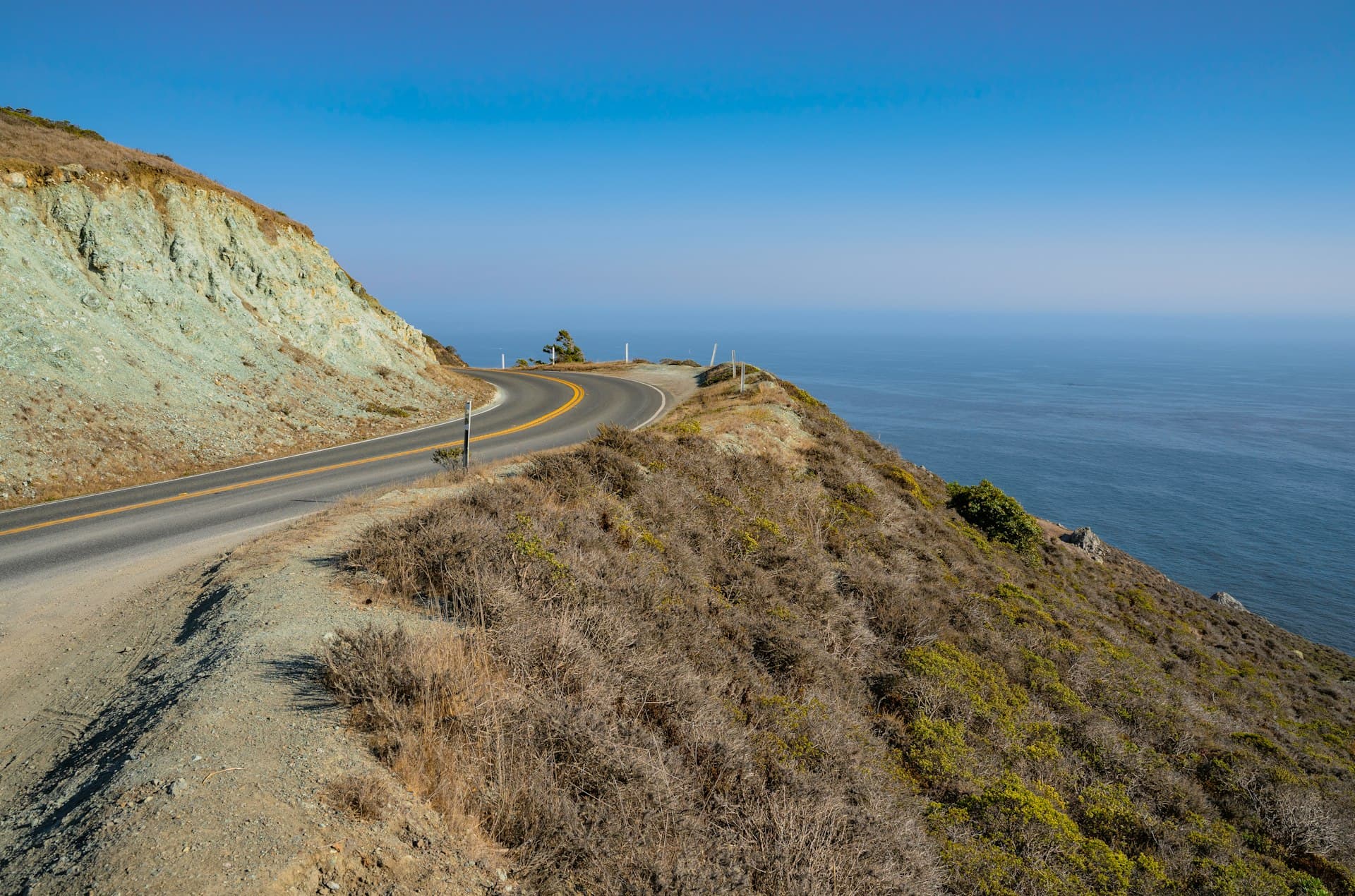 Scenic coastal highway with yellow center line curving along ocean cliffs under clear blue sky