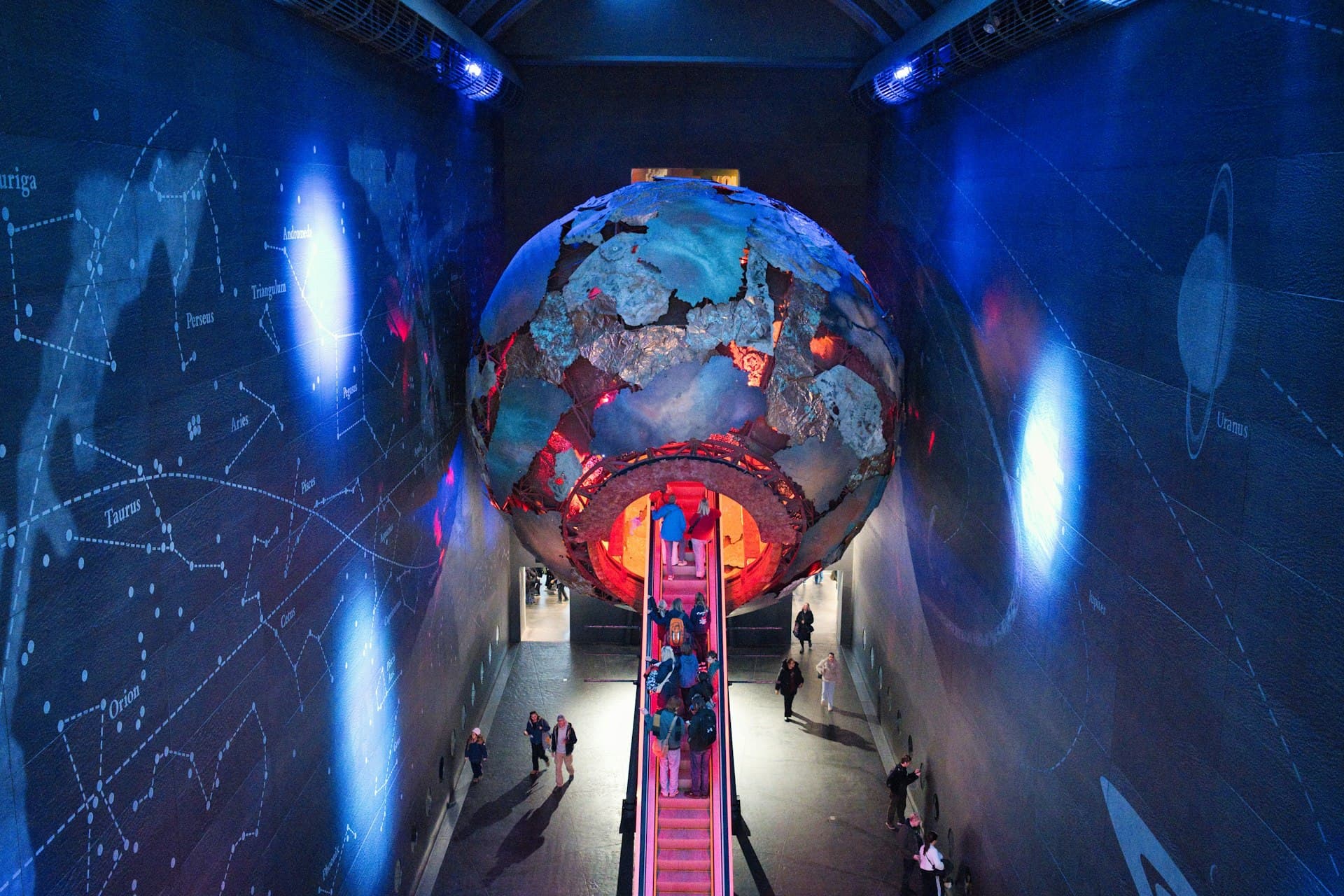 Families explore an illuminated interactive Earth globe inside a high-tech museum with constellation displays and red stairways in a blue-lit modern gallery space.