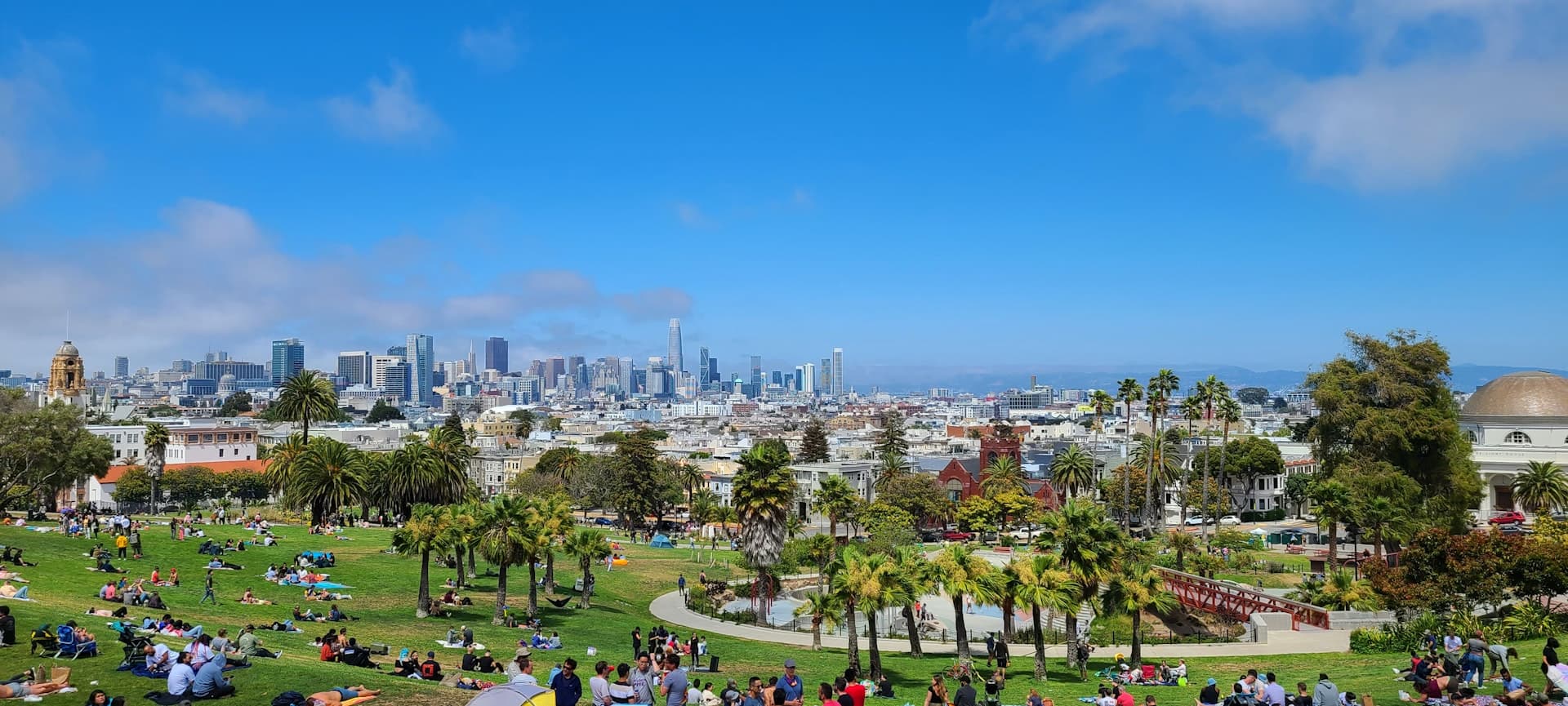 Families and groups enjoying a sunny day in an urban park with a city skyline backdrop, sitting on the grass and relaxing.