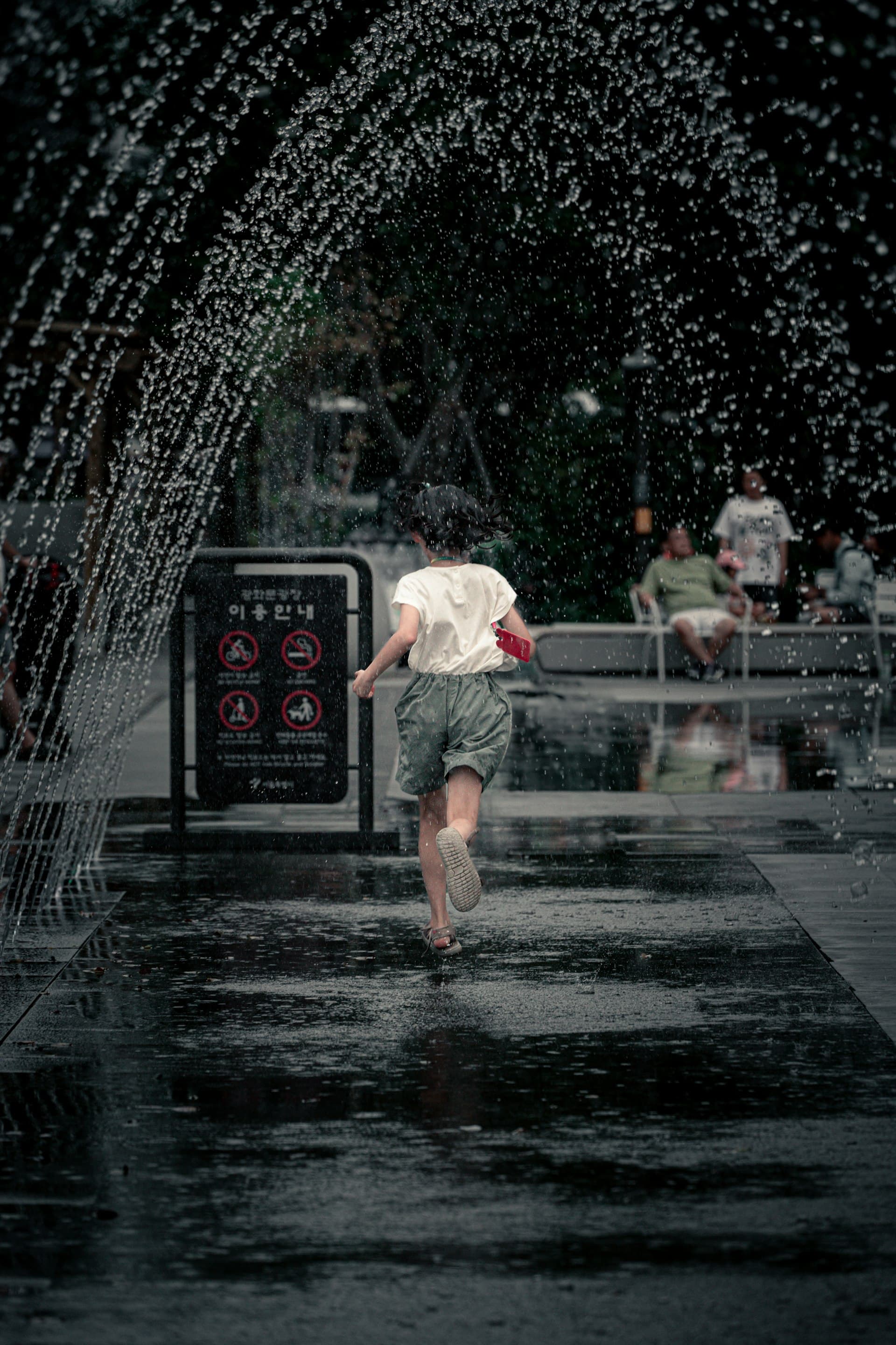 A school-age child runs through an arching water fountain on a rainy day, enjoying active outdoor play in an urban plaza.
