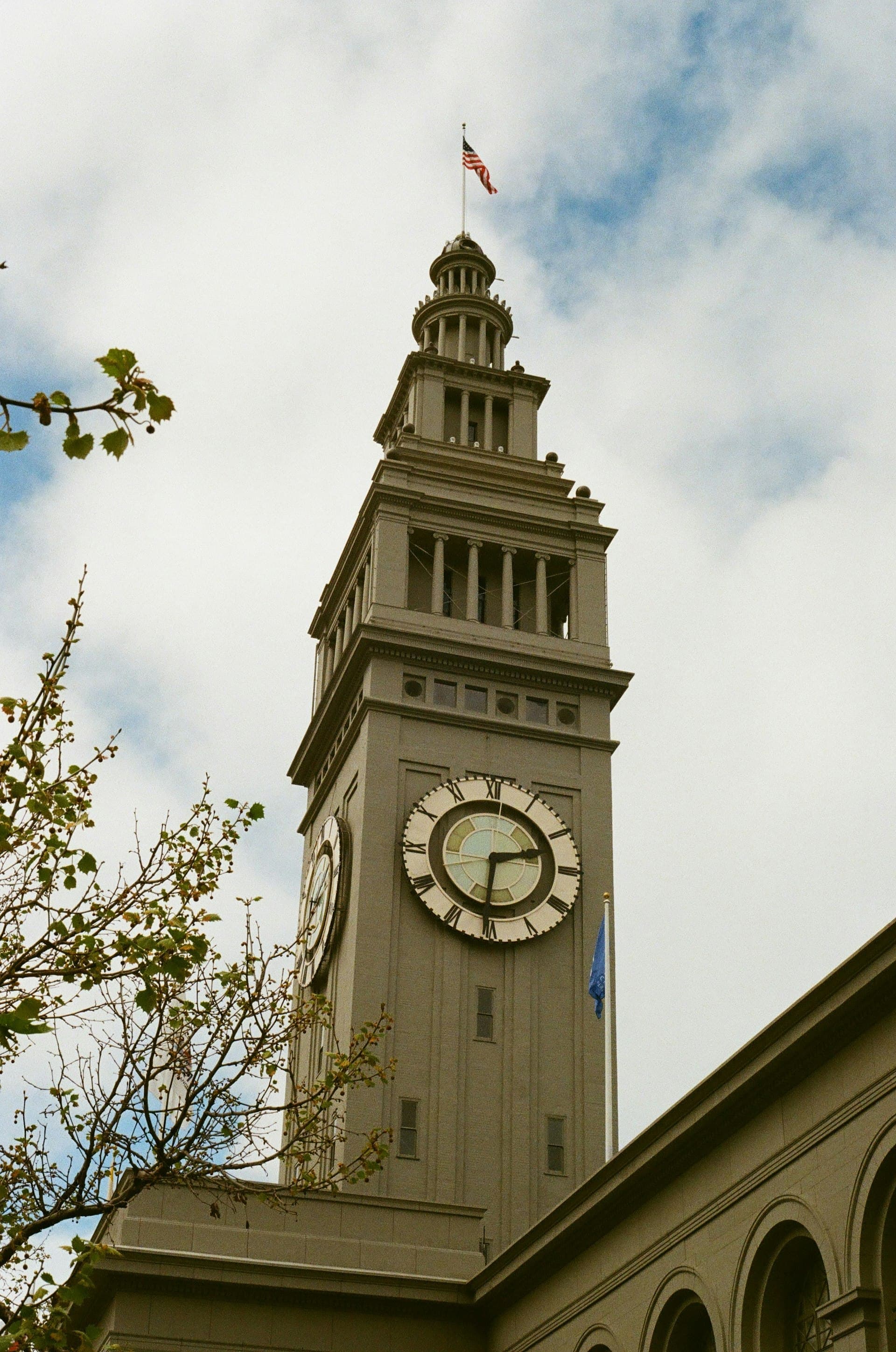 Tall historic clock tower with classical architecture and American flag against blue sky with tree branches in foreground.