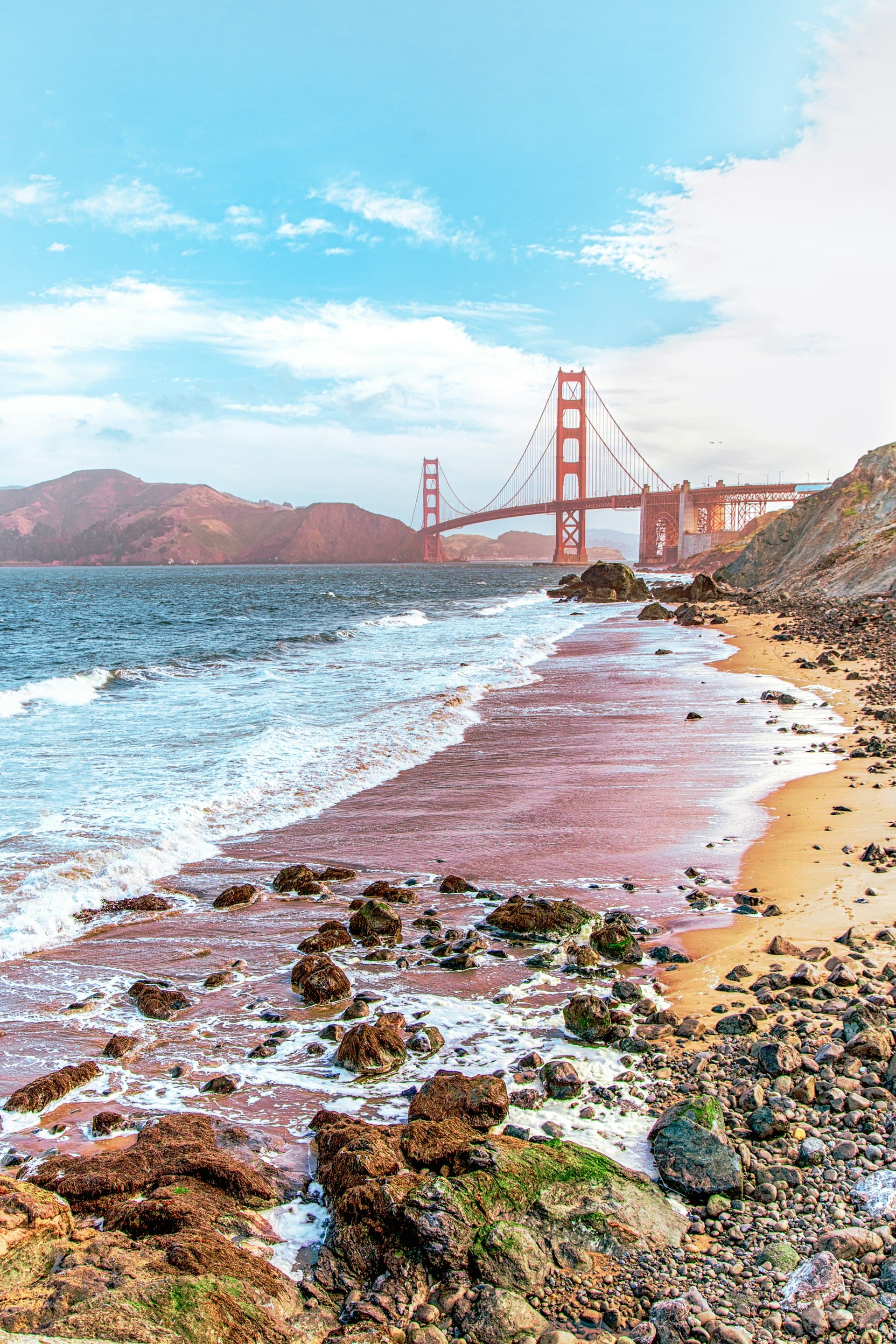 Golden Gate Bridge viewed from a rocky beach with turquoise ocean waves and sandy shore under blue sky