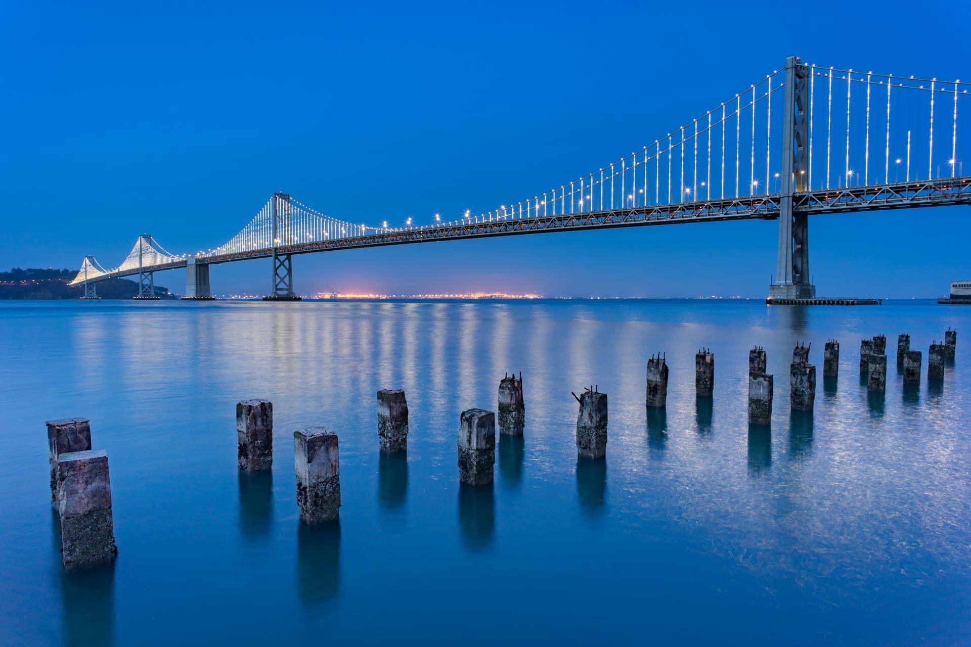 Bay Bridge lit up at night with old pilings in the foreground and city skyline glowing in the distance under a deep blue twilight sky.