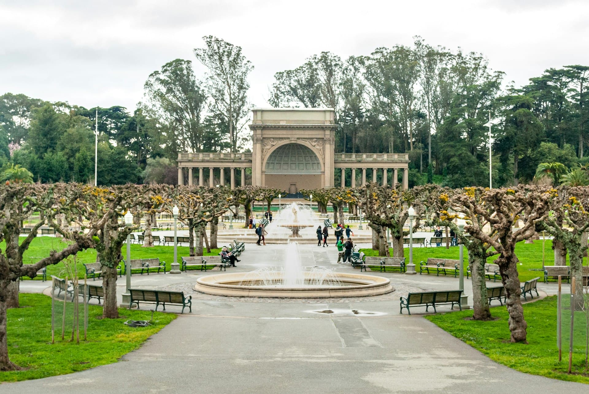 A grand urban park with a classical pavilion, manicured lawns, and visitors walking along tree-lined pathways with fountains in the foreground.
