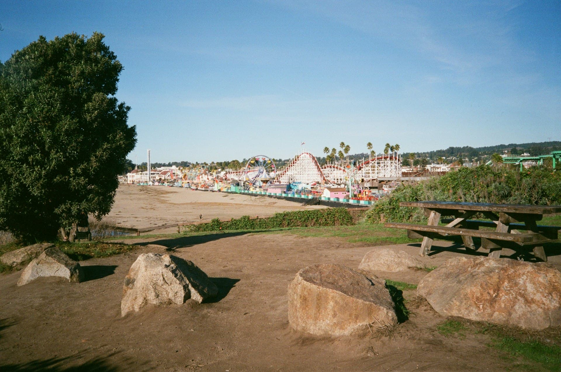 Amusement park with white roller coasters viewed from a sandy beach with picnic tables and rocks in foreground under clear blue sky.