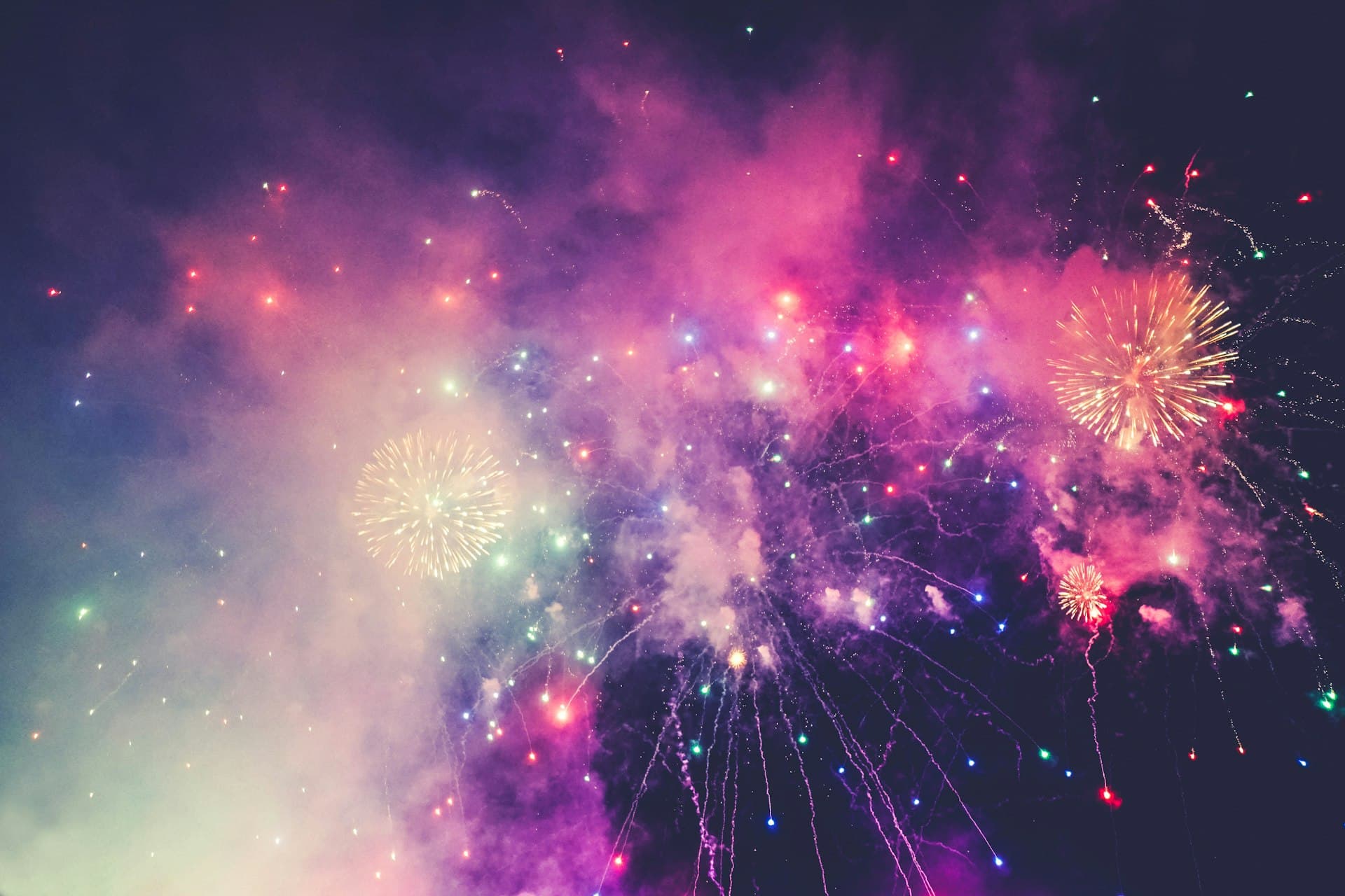 Colorful fireworks bursting across a dark night sky in shades of pink, purple, gold, and blue with sparkling smoke trails.
