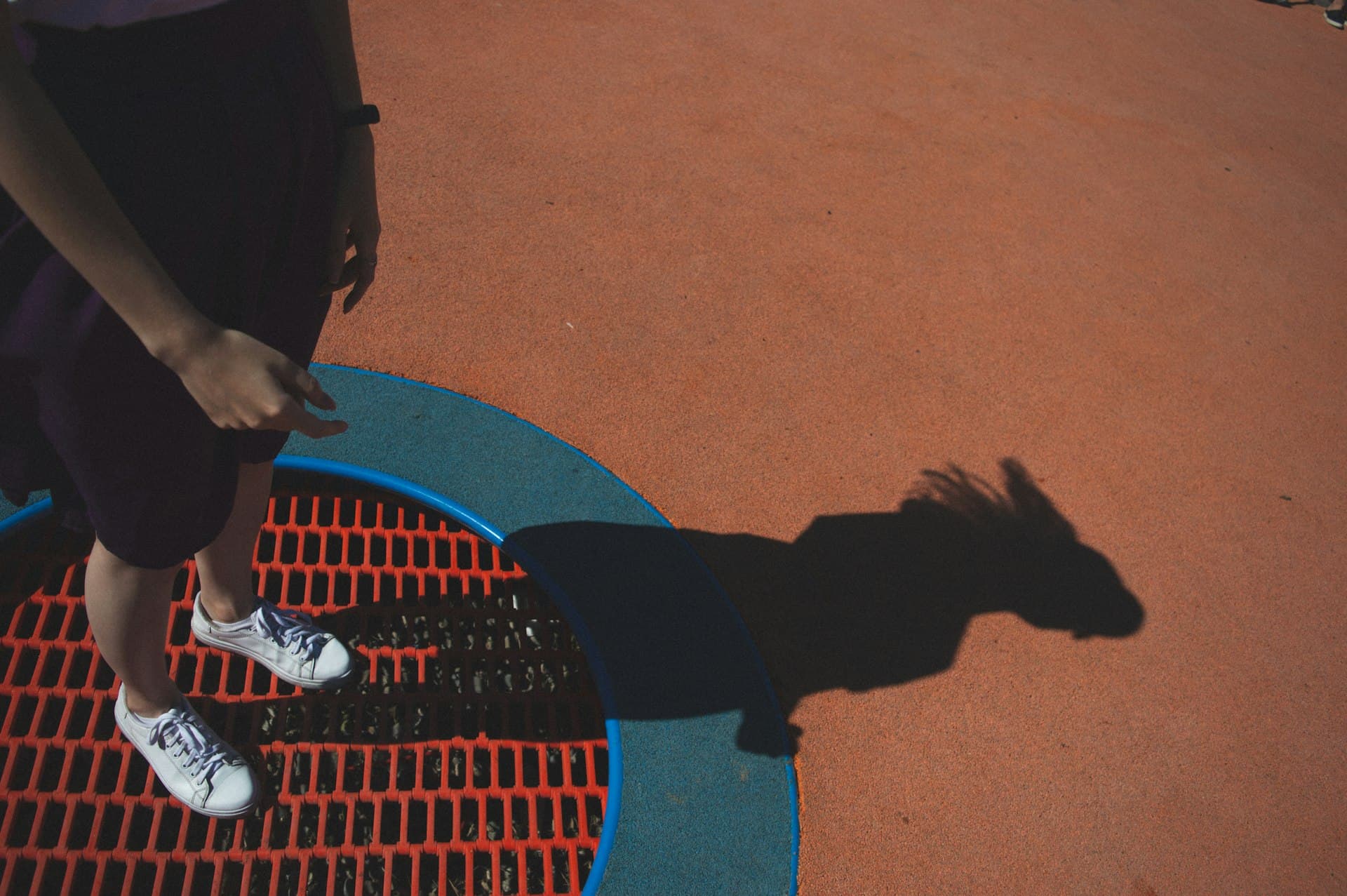 Child standing on a colorful playground mat outdoors, wearing white sneakers with their shadow visible on the ground.