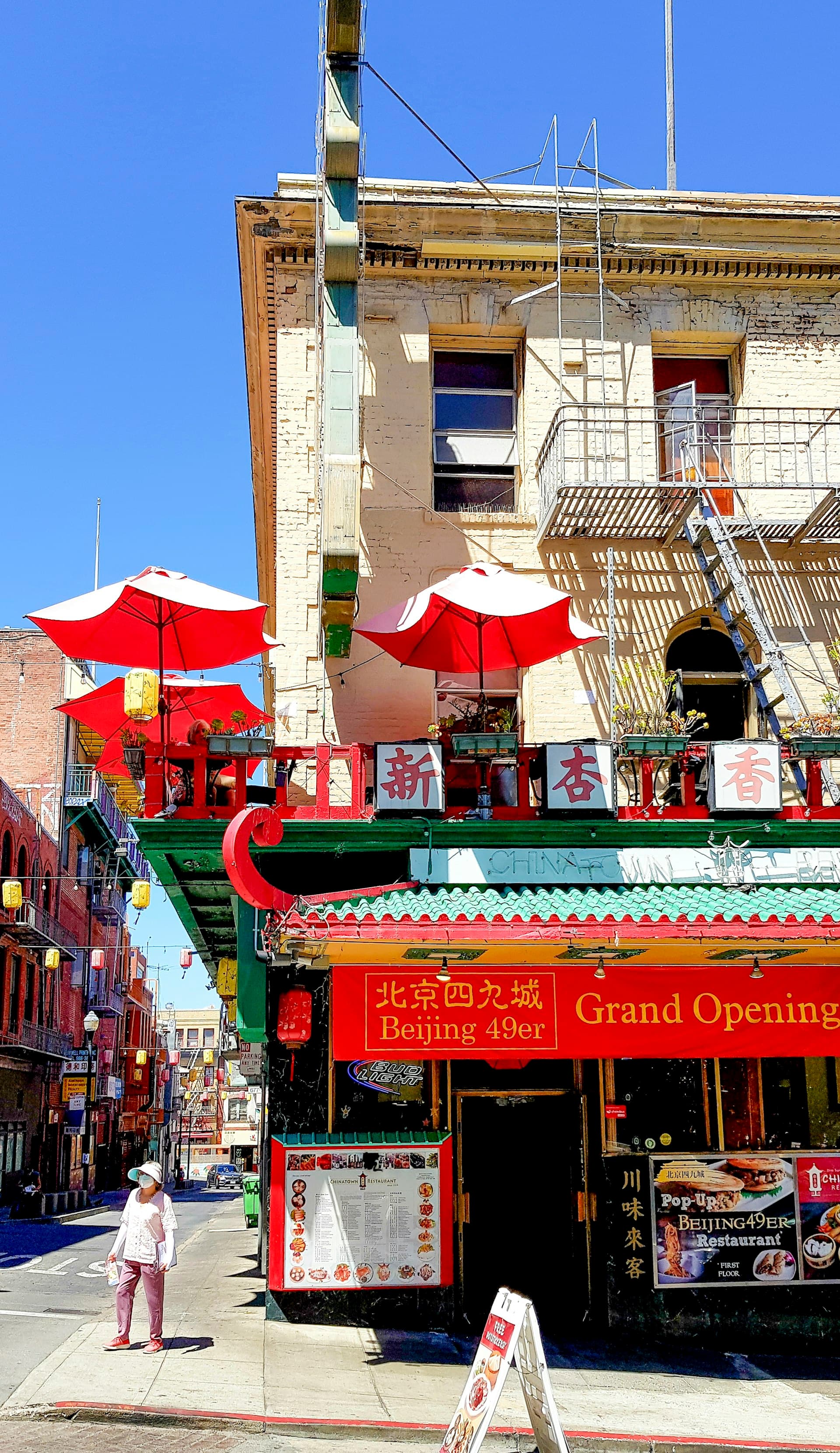 Person walking past a red and green Chinese restaurant with umbrellas on a sunny Chinatown street