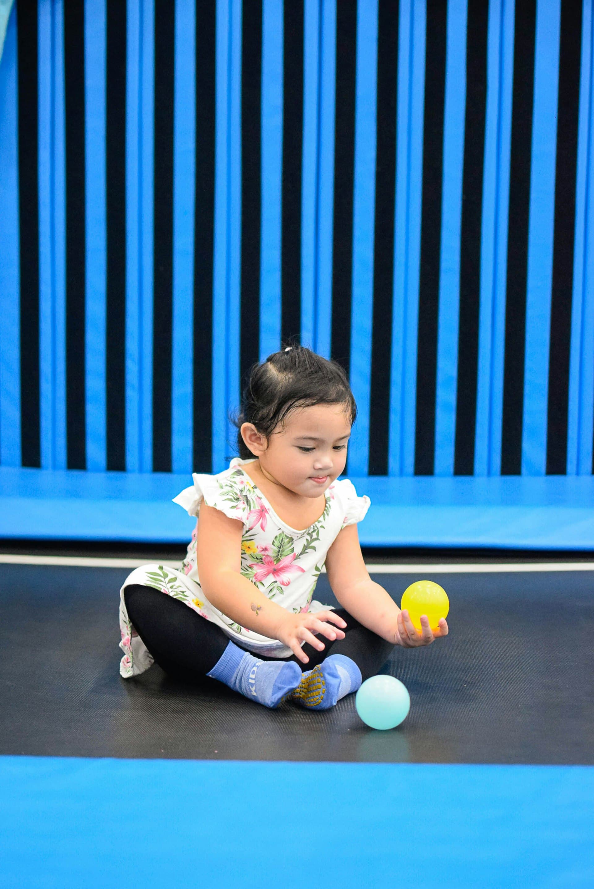 Young toddler sitting on a gym mat playing with yellow and light blue balls, wearing colorful clothing against a bright blue and black striped wall.
