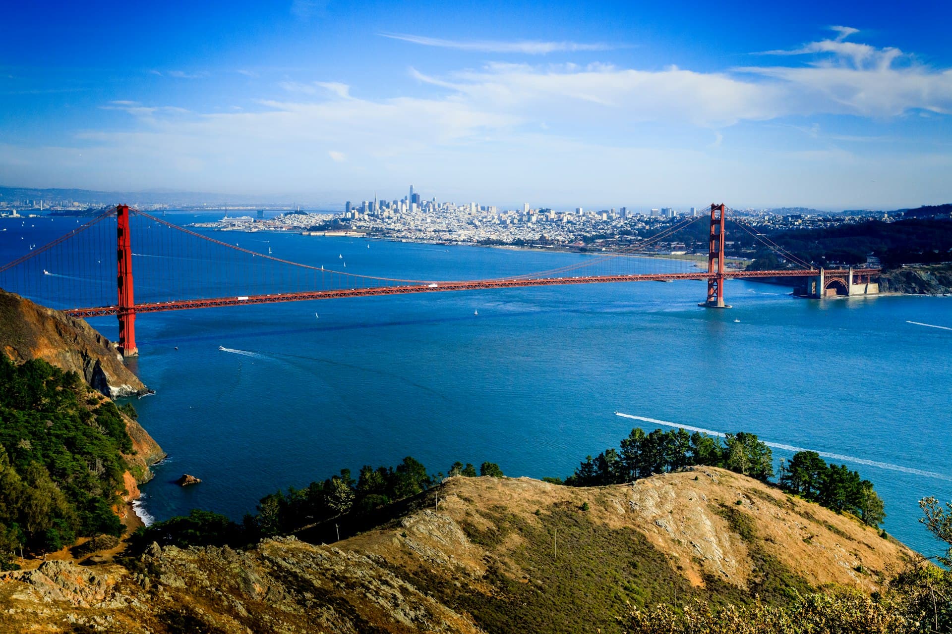 Aerial view of the Golden Gate Bridge spanning San Francisco Bay with city skyline and sailboats visible under clear blue sky