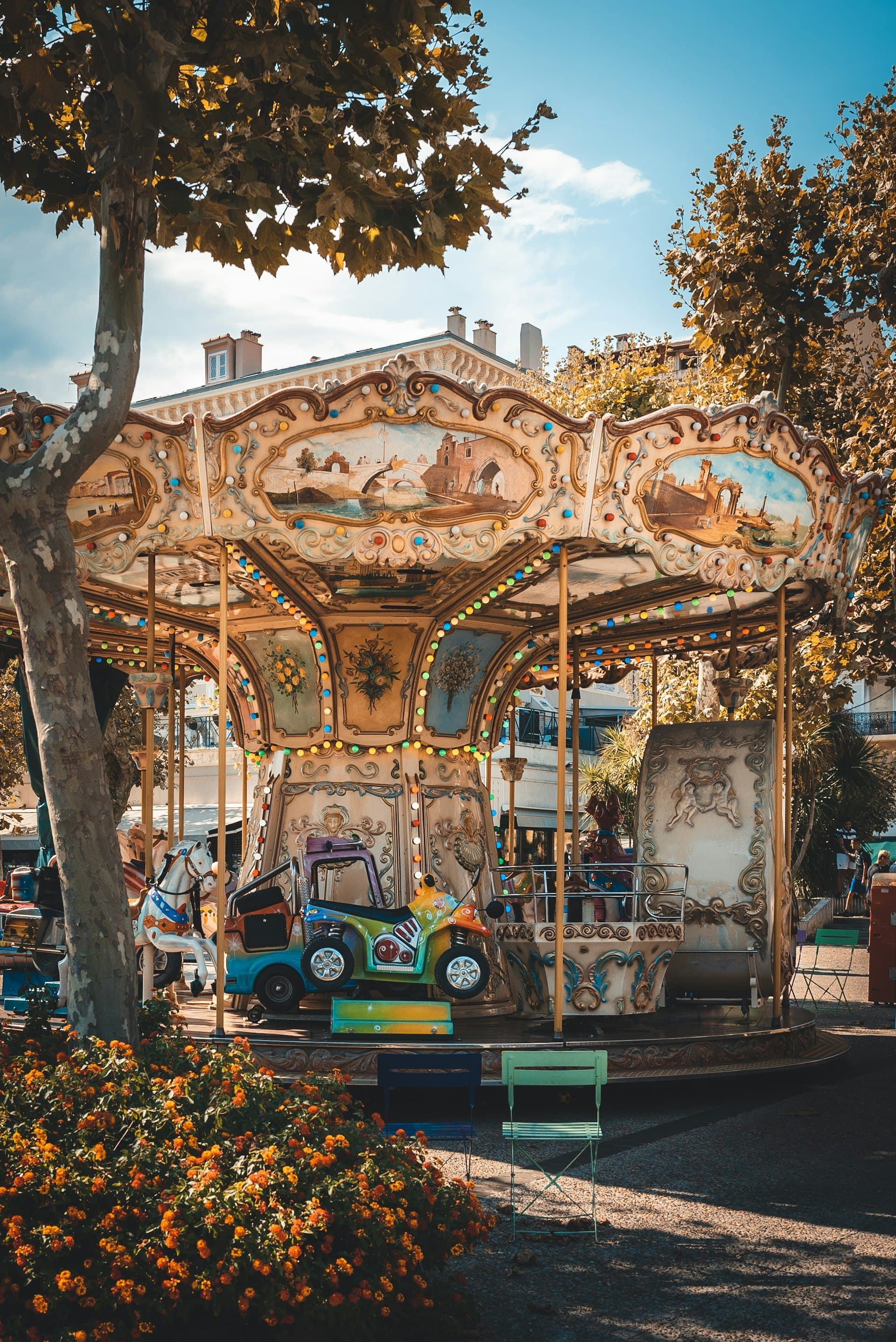 A vintage carousel with ornate wooden panels and colorful decorations in an autumn park setting, surrounded by golden trees and orange flowers.