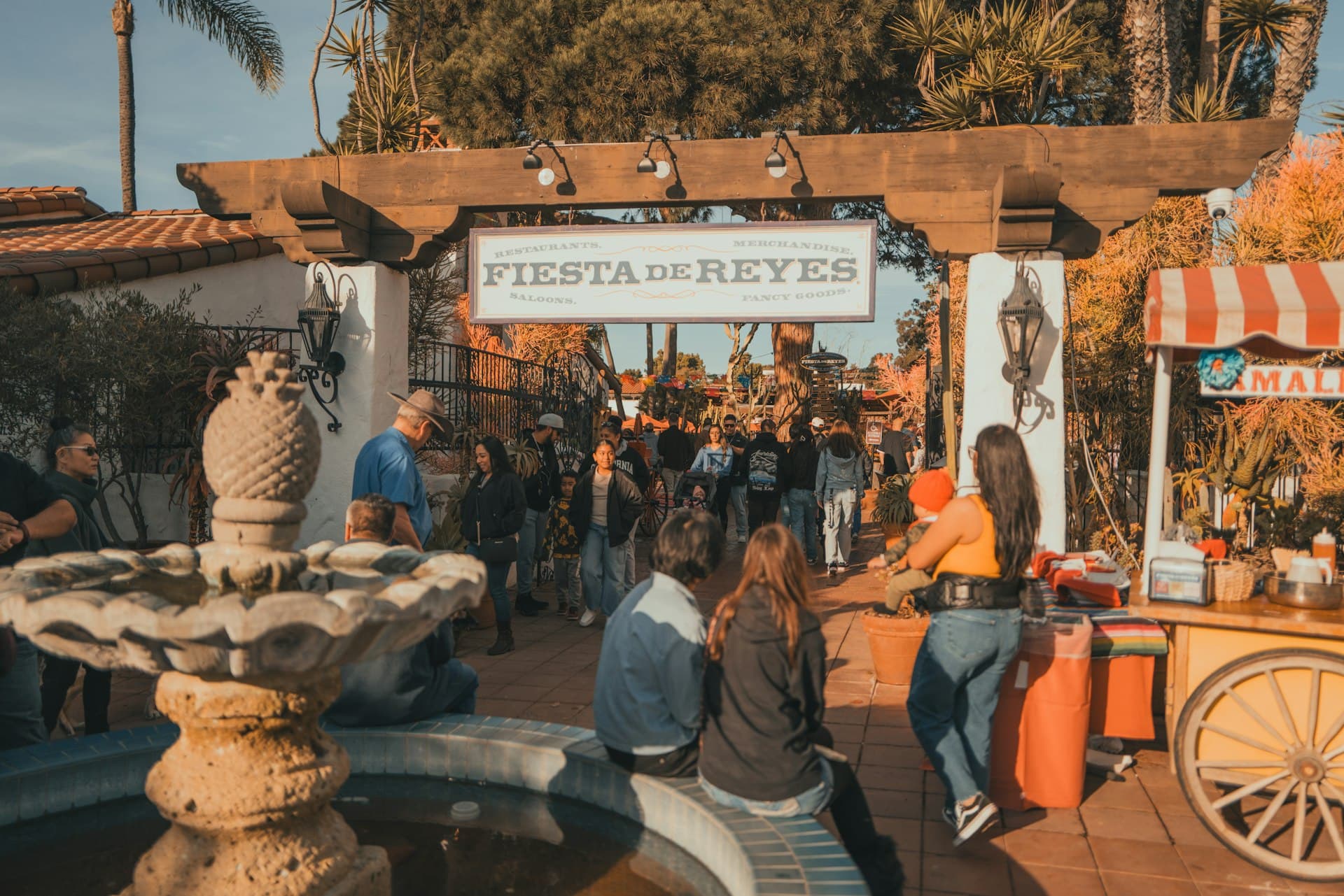 Family-friendly festival marketplace during golden hour with visitors exploring vendor stalls under wooden pergolas and palm trees.