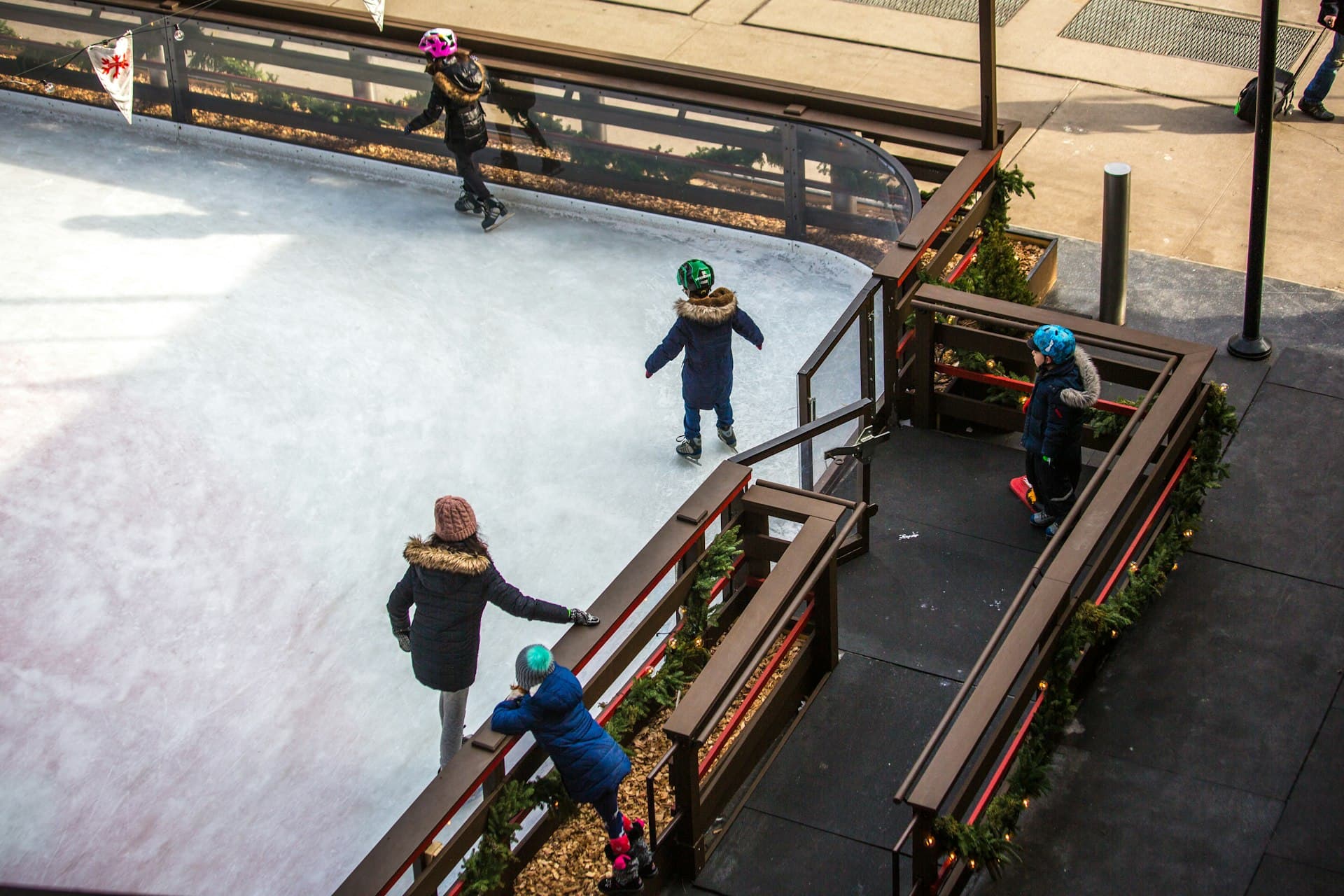 Children and parents enjoying ice skating together at an outdoor urban ice rink decorated with winter greenery.