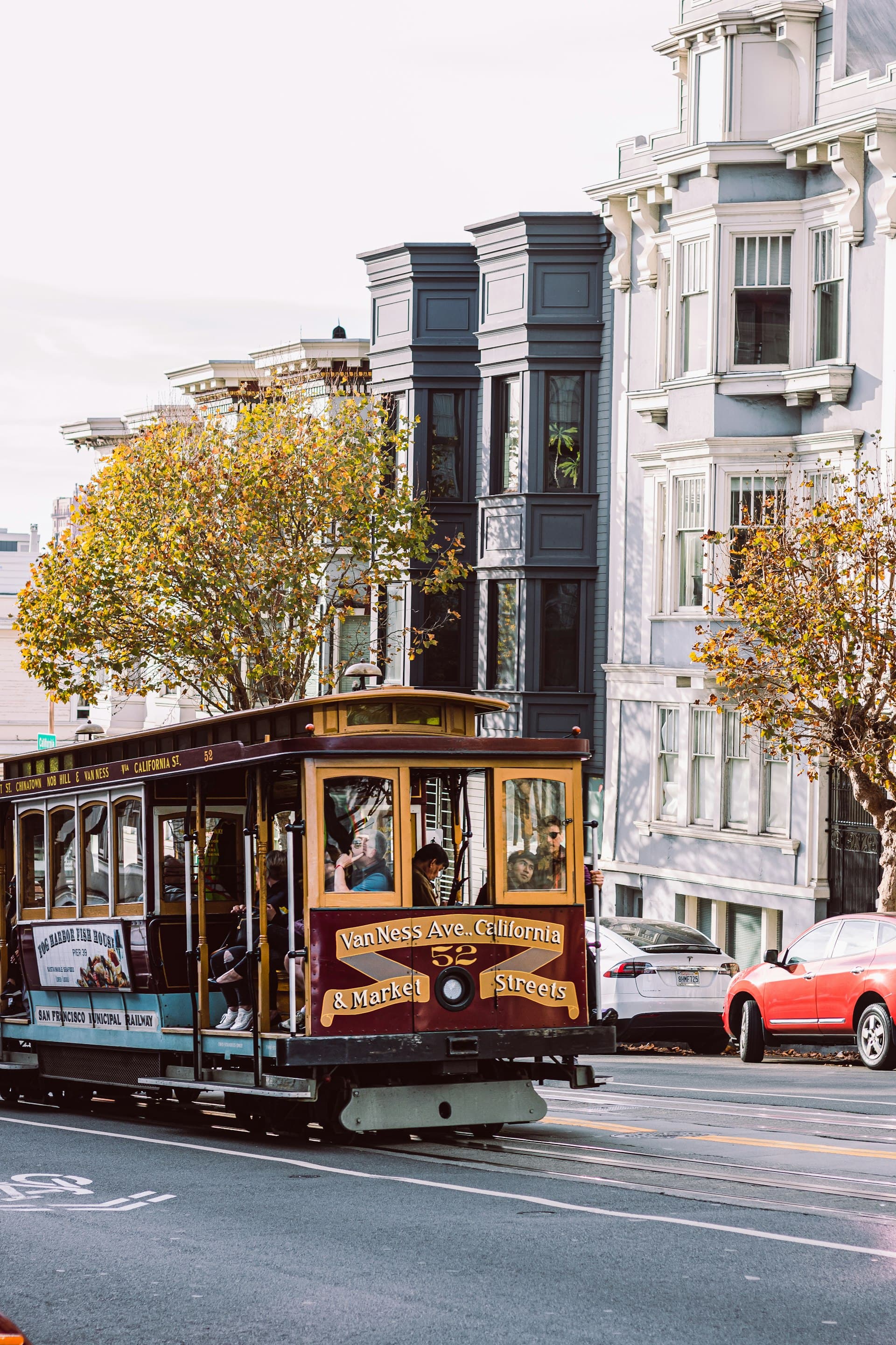 Historic San Francisco cable car with passengers traveling past Victorian buildings with fall-colored trees on Van Ness Avenue.