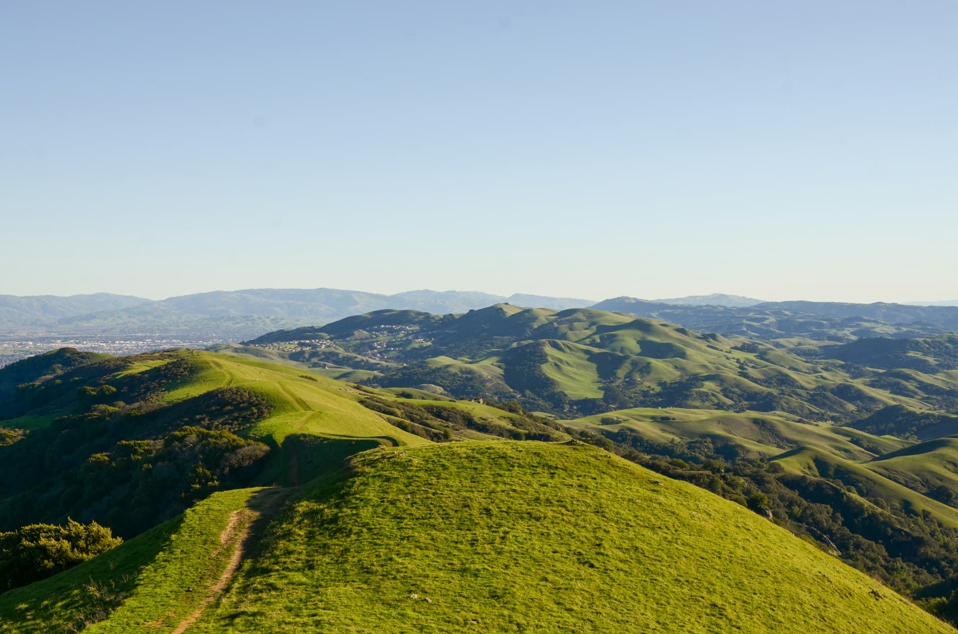 Wide aerial view of green rolling hills with a hiking trail running across lush grassland, surrounded by forested ridges and distant mountains under clear blue sky.