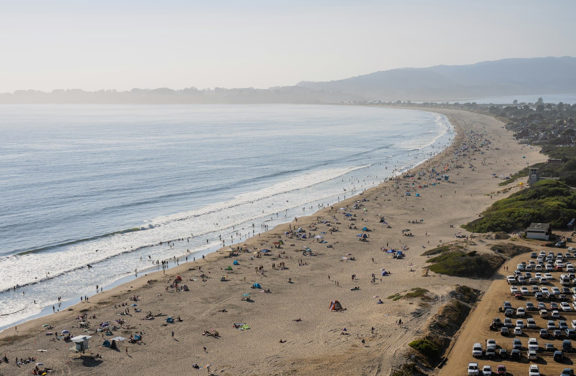 Aerial view of a busy beach with families swimming, wading, and relaxing on the sand, backed by mountains and coastal residential areas on a sunny day.