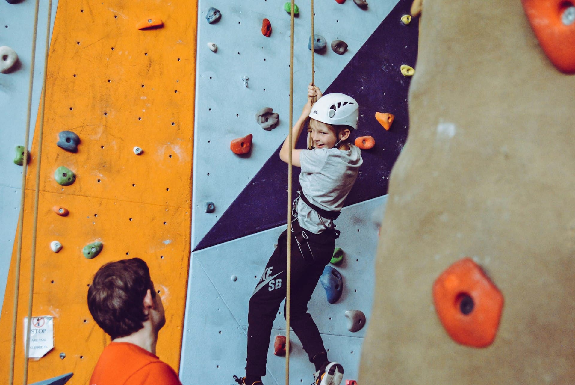 Child in white helmet climbing a colorful indoor rock wall with various handholds while another child watches below.