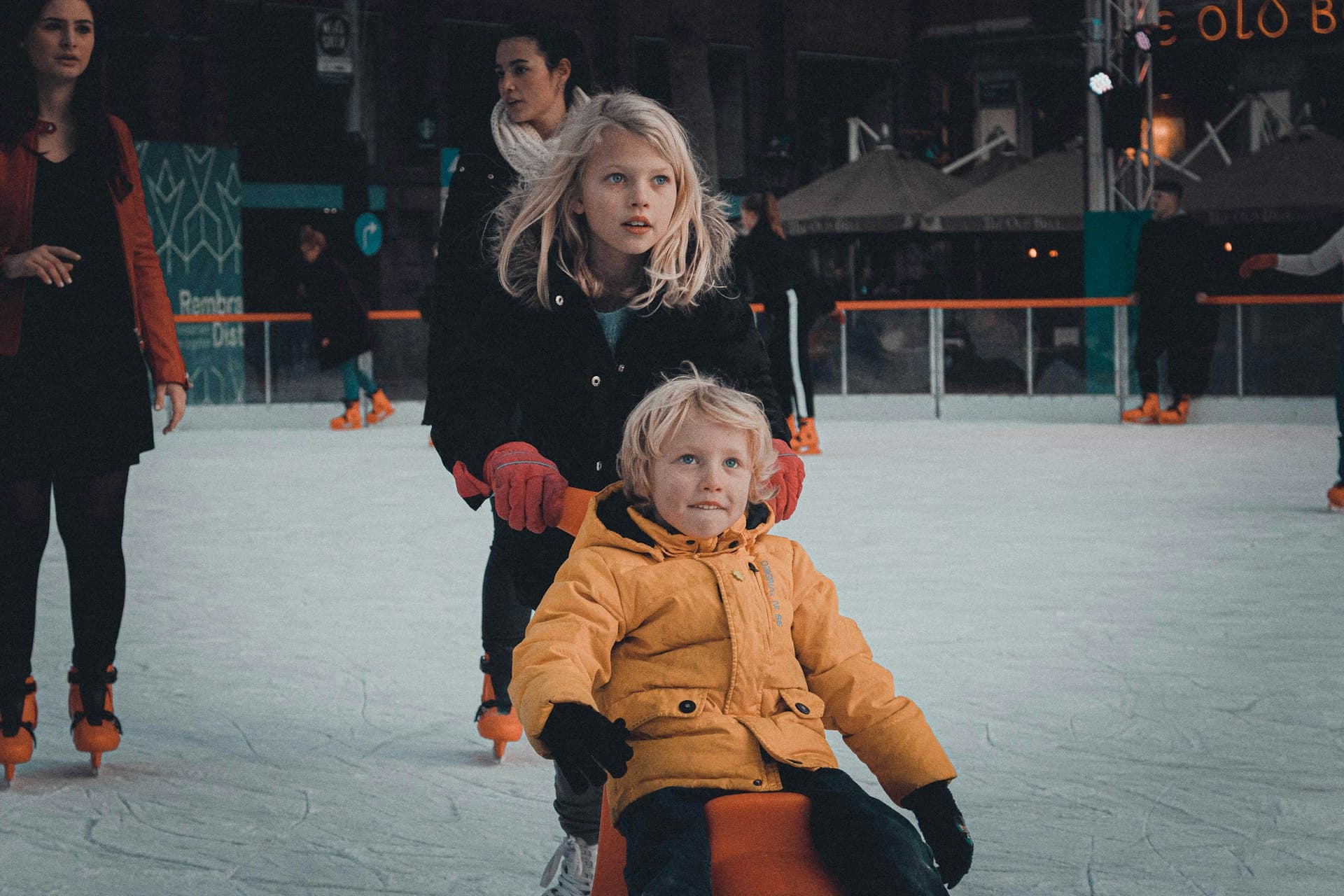 A young child in a yellow winter coat ice skates outdoors at a city rink, held by an older sibling wearing black, with other families skating in the background under evening lights.