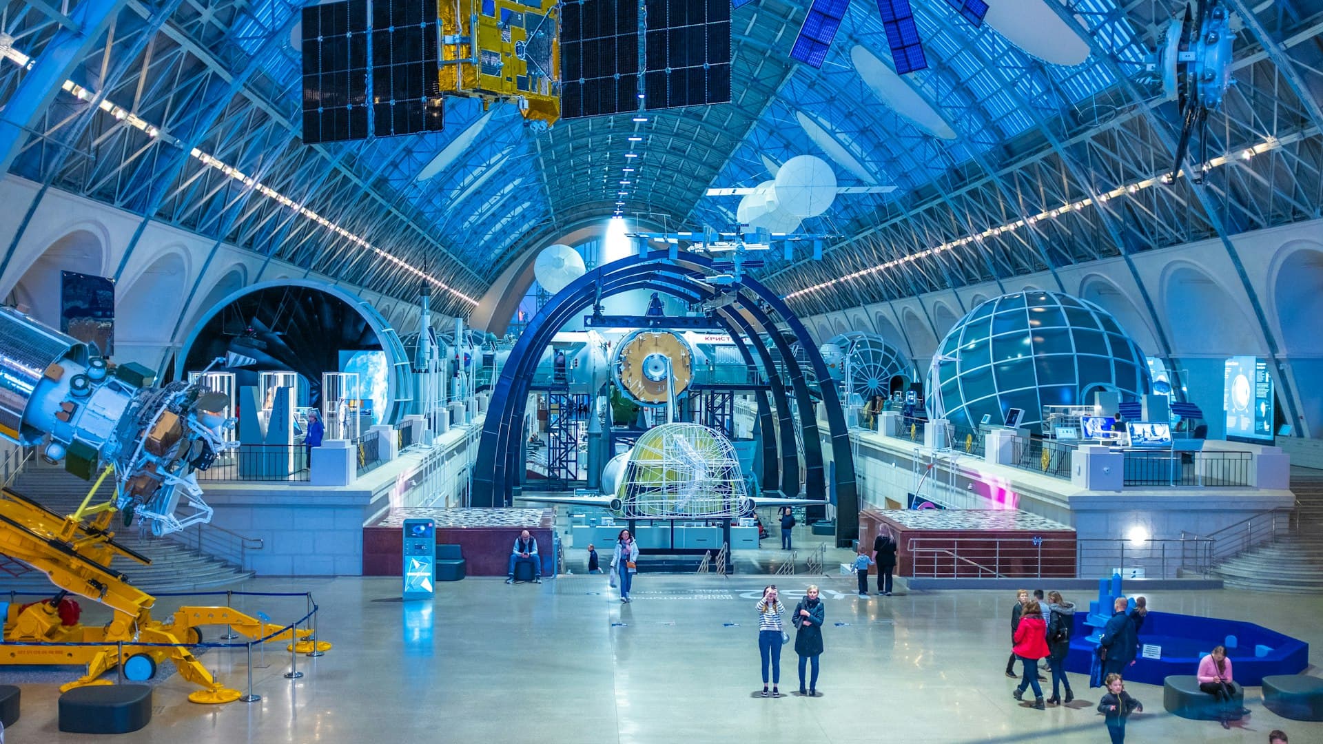 Families and children exploring a modern science museum with blue-lit architecture, satellite displays, and interactive exhibits in a large open hall.
